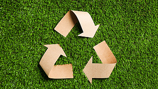 A recycling symbol made of brown cardboard lies on a green, artificial grass base. The arrows of the symbol are visible and give the impression of sustainability.