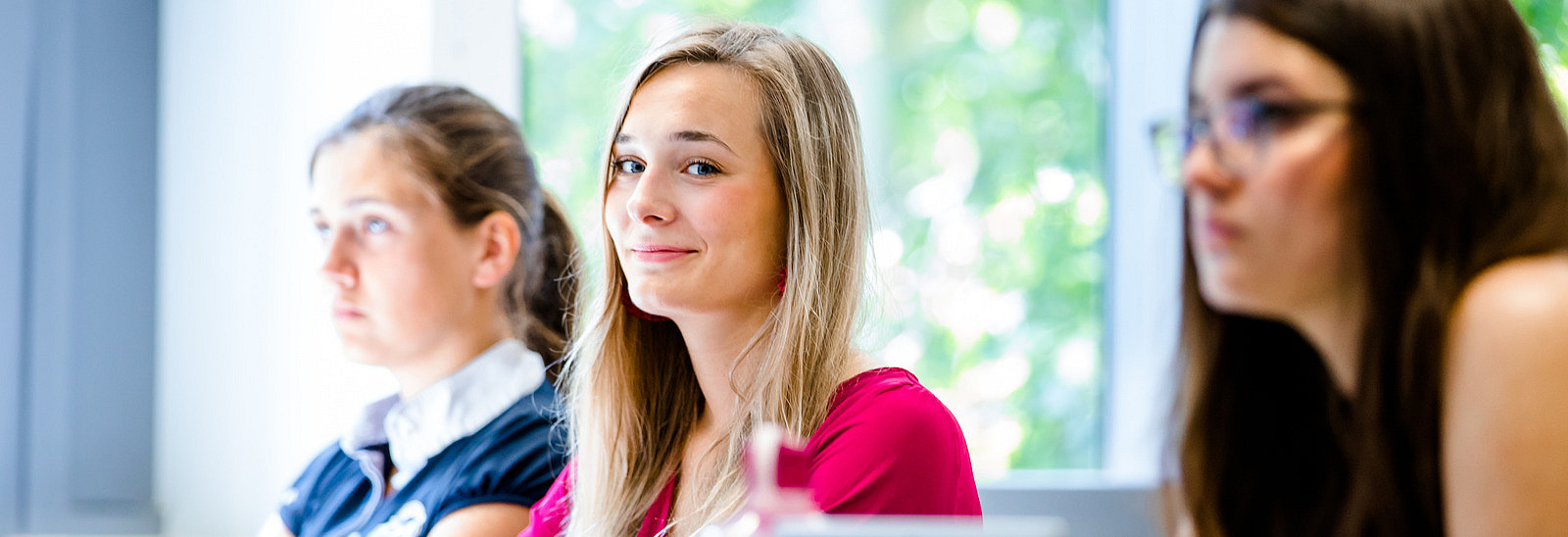 Female students in a row of seats in a seminar room. One of them smiles at the camera.