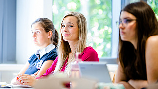 Female students in a row of seats in a seminar room. One of them smiles at the camera.