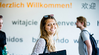 A young woman with long, wavy hair smiles at the camera while holding a black folder in her hand. In the background, the wall features the text "herzlich Willkommen!" and greetings in various languages. 