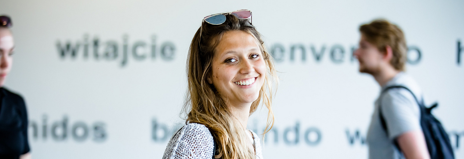 A young woman with long, wavy hair smiles at the camera while holding a black folder in her hand. In the background, the wall features the text "herzlich Willkommen!" and greetings in various languages. 