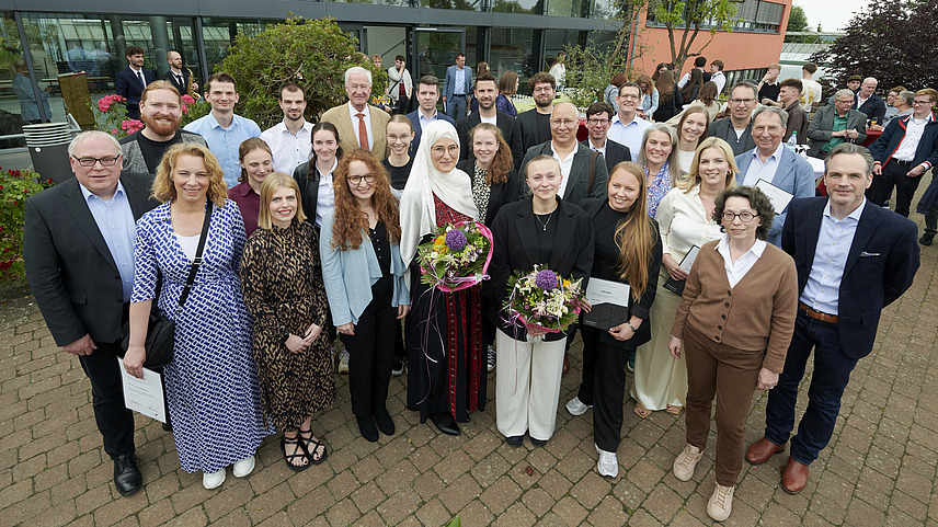 A large group of people smiles for a photo in front of a modern building with large windows. Some individuals hold bouquets in their hands. In the background, trees and plants are visible, and other guests stand casually.