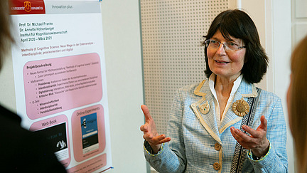 Eine Frau mit langen, dunklen Haaren trägt eine Brille und ein hellblaues Jackett mit, während ein Poster mit Informationen über ein Projekt im Hintergrund zu sehen ist.