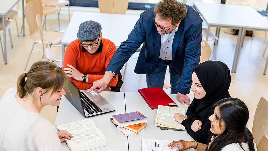 Fünf Personen sitzen an einem Tisch im Kreis und lesen gemeinsam in Büchern und auf einem Laptop. Eine Person trägt ein Kopftuch.