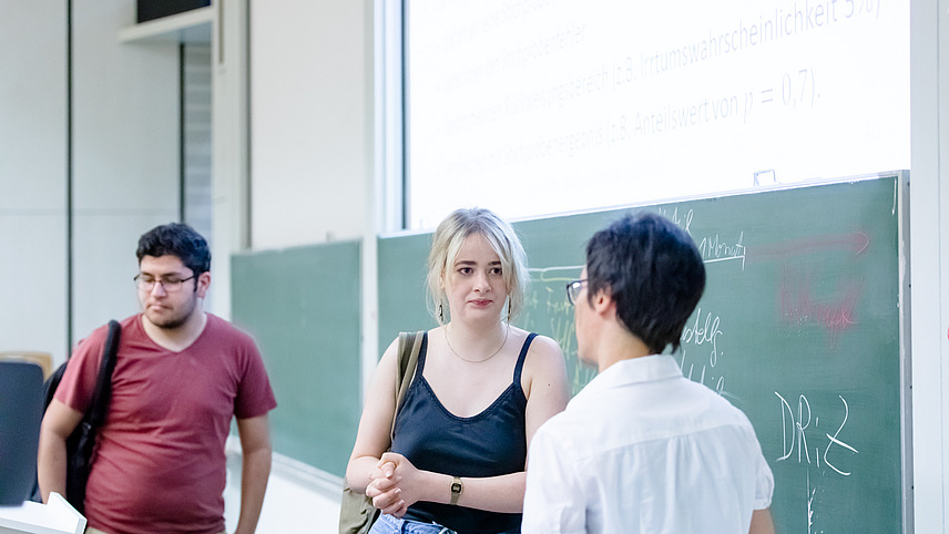 Eine Studentin in einem Vorlesungssaal redet mit der Dozentin vor der Tafel. Ein Student wartet hinter ihr.