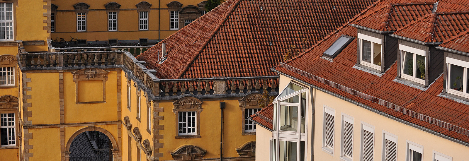 A view of the roofs of two buildings. The foreground building has yellow walls and a roof with red tiles. In the background, there are more yellow walls with windows visible. A row of bicycles is seen on the balcony.