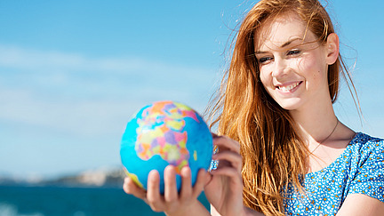 A red-haired young woman smiles as she holds a globe in her hand and takes a closer look at it. It is summer weather.