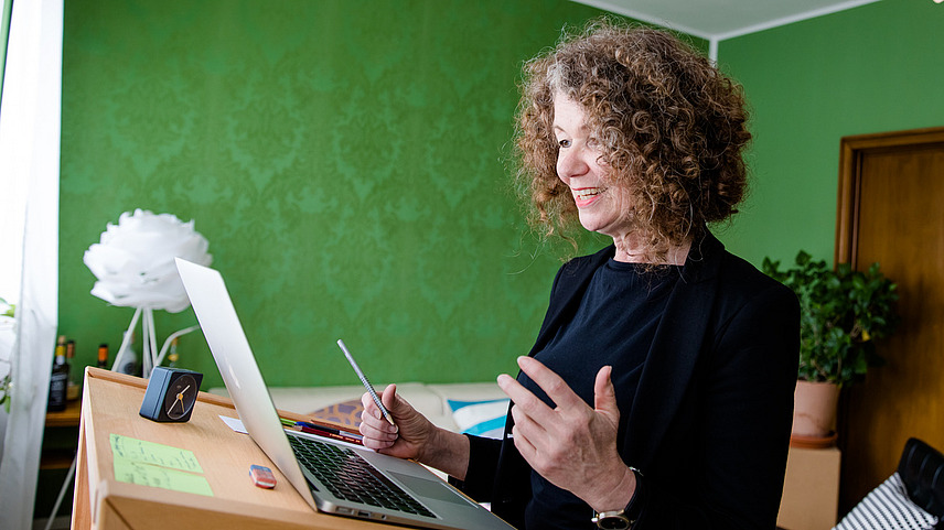 Eine Frau mit lockigem Haar steht an einem Pult und arbeitet an einem Laptop. Sie lächelt und spricht, während sie einen Stift in der Hand hält. Der Raum hat grüne Wände und einige Pflanzen sind im Hintergrund sichtbar.