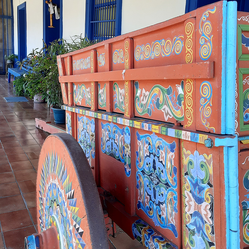 A decorative oxcart positioned on a tiled floor along a corridor. In the background, there are blue doors and windows, along with potted plants. 