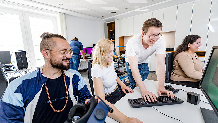 A few students are sitting or standing at computers. One student is holding a VR headset.