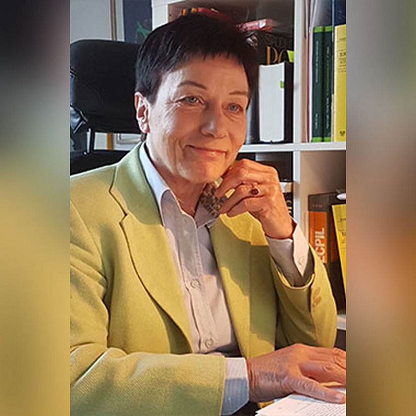 Eine Person mit schwarzem kurzen Haar und gelbem Blazer sitzt an einem Schreibtisch und lächelt in die Kamera