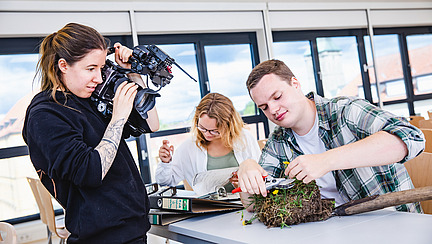 A young woman dressed in black uses a large camera to photograph a piece of excavated meadow, which is being positioned on a table in front of her by a man. In the background, another woman is reading a file folder.