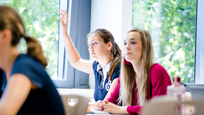 Zwei Studentinnen an einem Tisch in einem Seminarraum an einem sonnigen Tag. Eine von ihnen hebt die Hand zur Meldung.