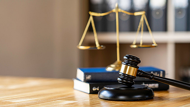 The picture shows a court symbol, consisting of a judge's gavel and a pair of scales, which stands for justice. Several books can be seen in the background, which refer to legal topics.