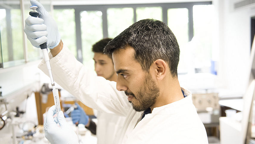 Two men in a laboratory fill liquids into a plastic tube using pipettes.