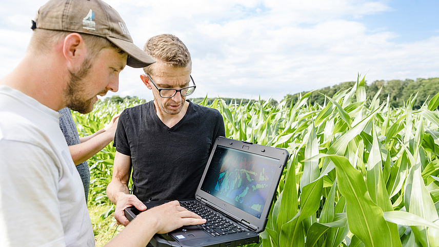 Two people standing in a field of tall, green plants (presumably corn). One of the people is holding a laptop, while the other person appears to be looking at the laptop and pointing at it. In the background are other plants