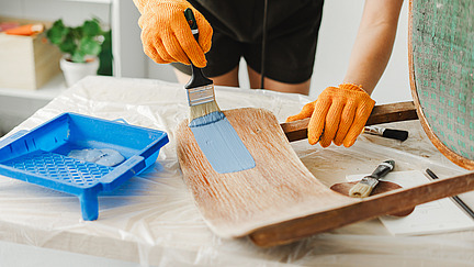 A person wearing orange gloves is painting blue paint onto a piece of wood with a brush. There is a blue paint tray next to her.