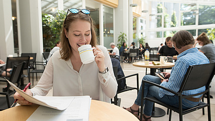 A smiling woman sits at a round wooden table in a light-colored café, holding a cup. In front of her are documents that she is leafing through. The room is decorated with plants and large windows, creating a pleasant atmosphere. Other guests can be seen in the background, sitting at tables and talking or working.