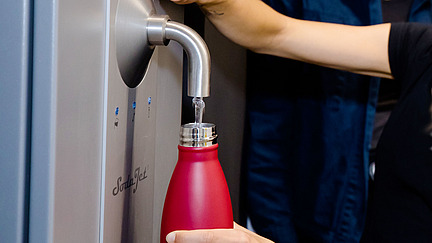 The picture shows a person holding a red water bottle to a water dispenser. Clear water is flowing into the bottle from the dispenser spout. 