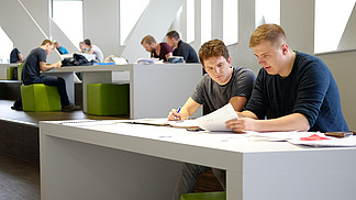 Two young men are sitting at a large table studying together. The man on the left is writing on a piece of paper while the man on the right is showing him documents. In the background, there are more people working at tables.