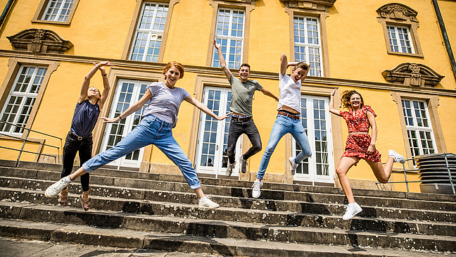 Fünf Studierende springen auf einer Treppe vor dem Schloss mit ausgebreiteten Armen in die Luft.