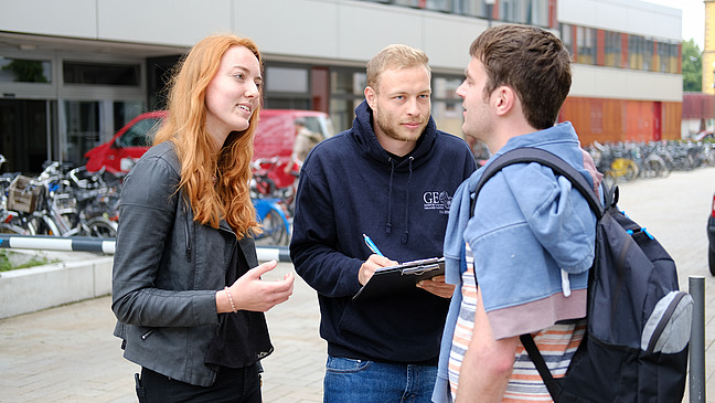 Three people in front of a building, one holding a clipboard and taking notes