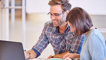 A man and a woman are sitting together at a desk, the mood is relaxed and they seem to enjoy working together. The man is working on his laptop and the woman is leafing through a book.