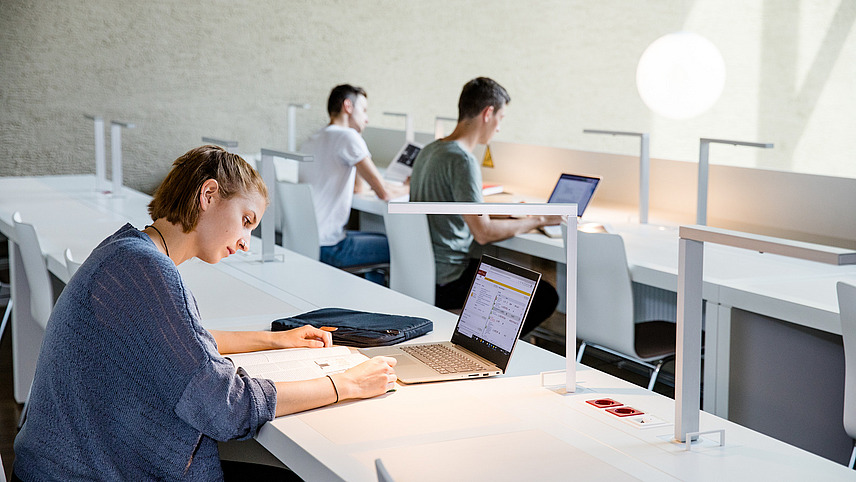 Drei Studierende sitzen an Tischen in der Bibliothek am Westerberg und lesen oder arbeiten am Laptop.