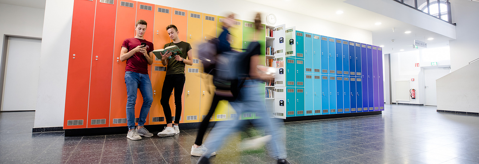 Zwei Studenten lehnen an Spinden, deren Farbreihenfolge einem Regenbogen nachempfunden ist.