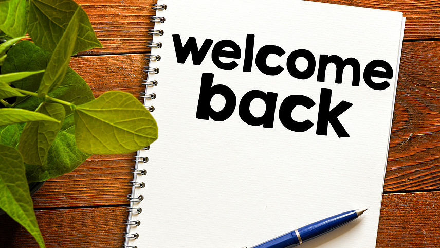 A blue pen lies on a white notepad with the inscription 'welcome back' on a wooden base. 