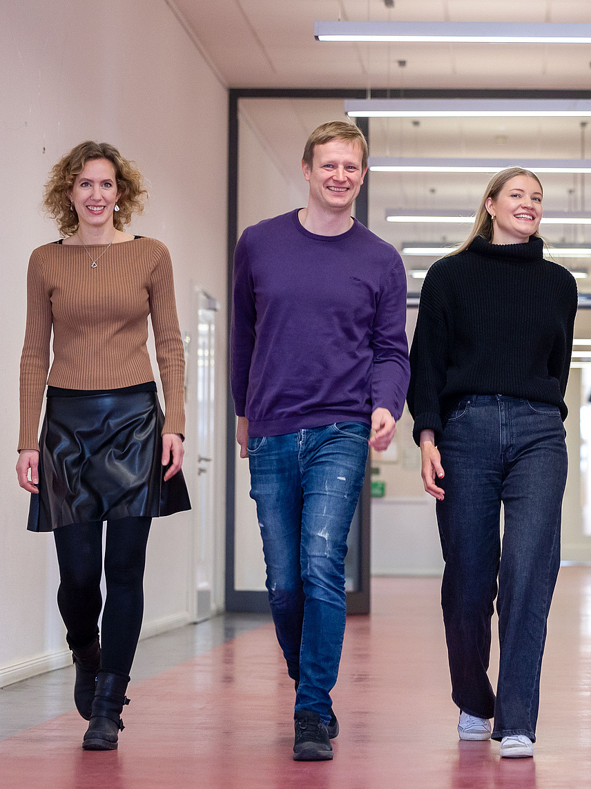 Five young adults walk together in a hallway towards the camera. The hallway has light-colored walls and a red floor