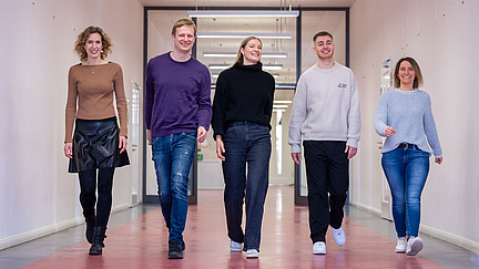 Five young adults walk together in a hallway towards the camera. The hallway has light-colored walls and a red floor