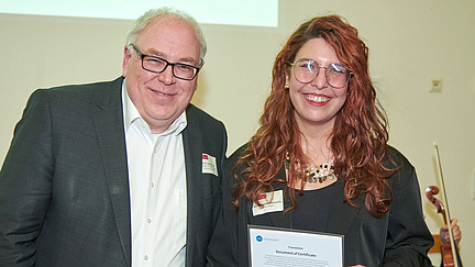 In a festive setting, two people stand next to each other, smiling at the camera. A woman holds a certificate in her hand, while a man stands beside her.