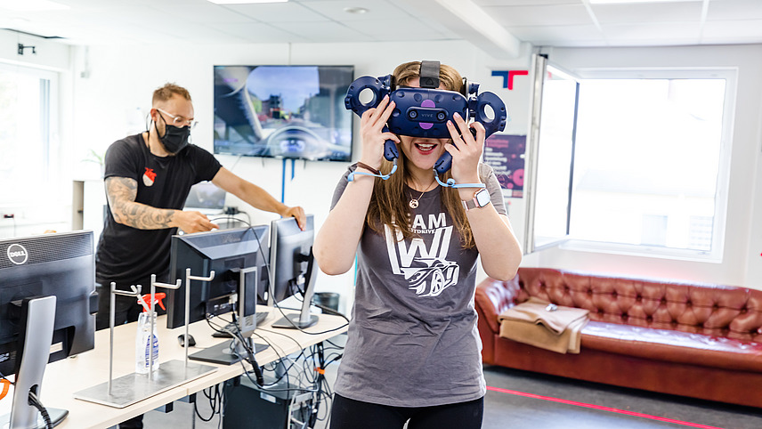 Im Vordergrund steht eine Frau, die eine VR Brille ausprobiert, während ein Mann im Hintergrund auf die Bildschrime vor ihm schaut.