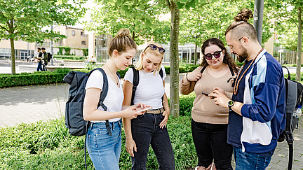 Four students look at their cell phones together. You can just make out the canteen in the background.