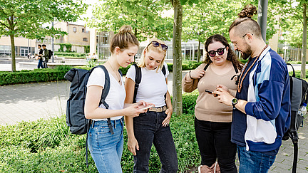 Vier Studenten schauen gemeinsam aufs Handy. Im Hintergrund ist gerade noch die Mensa zu erkennen.