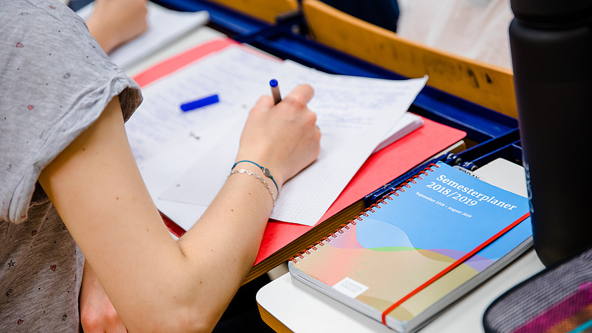 A student in a lecture hall takes notes on a piece of paper.