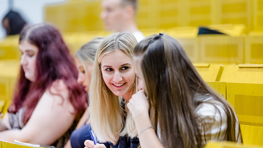 Zwei Studentinnen unterhalten sich im Vordergrund. Im Hintergrund sind weitere Studierende auf den gelben Sitzreihen eines Hörsaals zu sehen, die sich Notizen machen.