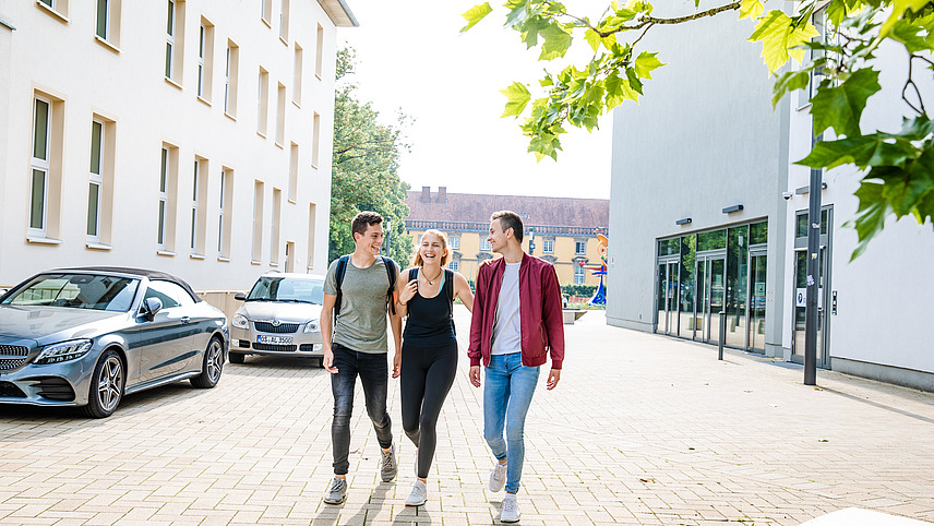 Three students are talking while walking outside on a sunny day. The castle can be seen in the background between two other buildings.