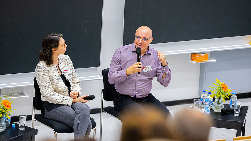 Two people sit on stage discussing in front of an audience.