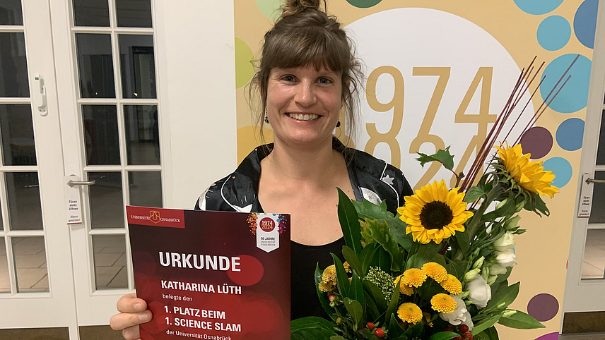 Das Bild zeigt eine junge Frau, die lächelt und eine Urkunde des Science Slam in der Hand hält. Sie hat einen Blumenstrauß mit Sonnenblumen und anderen Blumen in der anderen Hand. Im Hintergrund ist ein buntes Motiv mit dem Jahr "1974-2024" zu sehen, das auf den 50. Jahrestag der Universität Osnabrück hinweist.