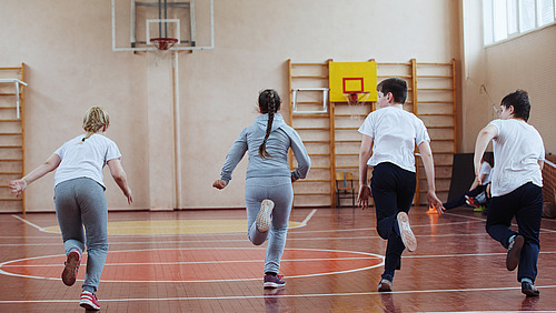 Primary school children a sport lesson indoors Vier Schülerinnen und Schüler rennen durch eine Turnhalle.
