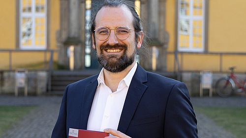 Prof. Zander stands in the inner courtyard of Osnabrück Castle. He holds a folder in his hand and smiles at the camera.