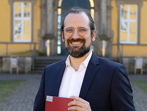 Prof. Zander steht im Innenhof des Osnabrücker Schlosses. Er hält eine Mappe in der Hand un lächelt in die Kamera.