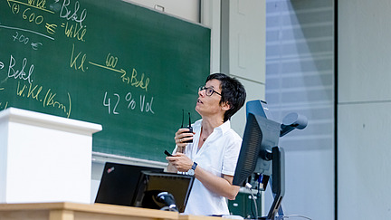 A lecturer is standing in a lecture hall giving a lecture. The blackboards in the background are full of notes.