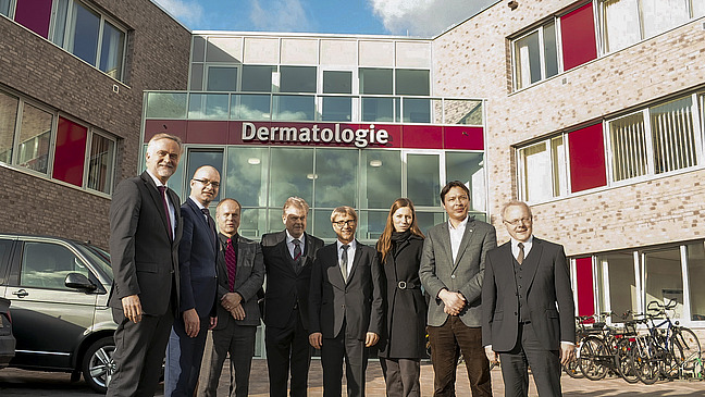 Eine Gruppe von acht Menschen steht lächelnd vor einem modernen Gebäude mit einer großen Beschriftung „Dermatologie“.