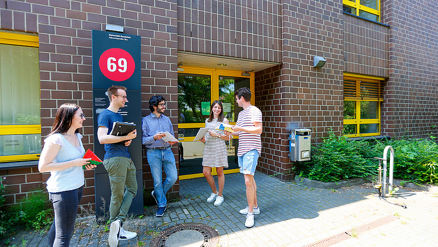 Fünf Studierende stehen bei gutem Wetter vor dem Eingang eines Backsteinhauses mit gelben Fenstern, das die Gebäudenummer 69 trägt. Dabei handelt es sich um das Mathematikgebäude der Universität. Alle fünf halten Arbeitsmaterialien in den Händen.