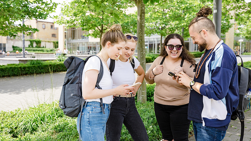 Vier Studenten schauen im Schatten eines Baumes gemeinsam auf ein Handy. Im Hintergrund ist das Mensagebäude zu erkennen.