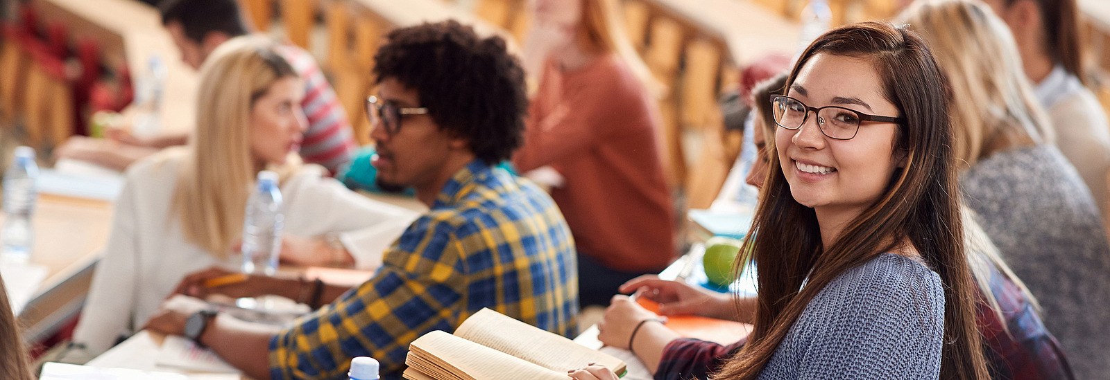 Eine junge Frau sitzt in einem Hörsaal mit einem Buch in der Hand und schaut in die Kamera. In den Sitzbänken um sie herum sitzen andere Studierende die sich unterhalten, schreiben oder nach vorne schauen. 