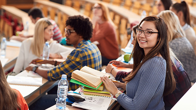 A young woman sits in a lecture hall with a book in her hand and looks into the camera. Other students are sitting in the benches around her, talking, writing or looking ahead.